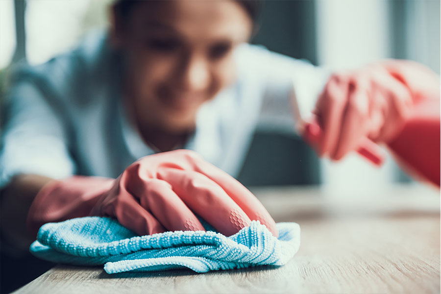 A cleaning woman in a blue cleaning uniform and pink gloves spraying a residential table with eco-friendly cleaning supplies and a blue washcloth