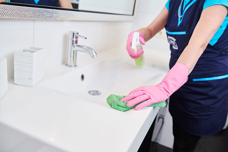 A maid in a blue uniform and pink gloves using a cleaning spray to wipe down a bathroom sink in a residential home in St. Charles and St. Louis, MO.