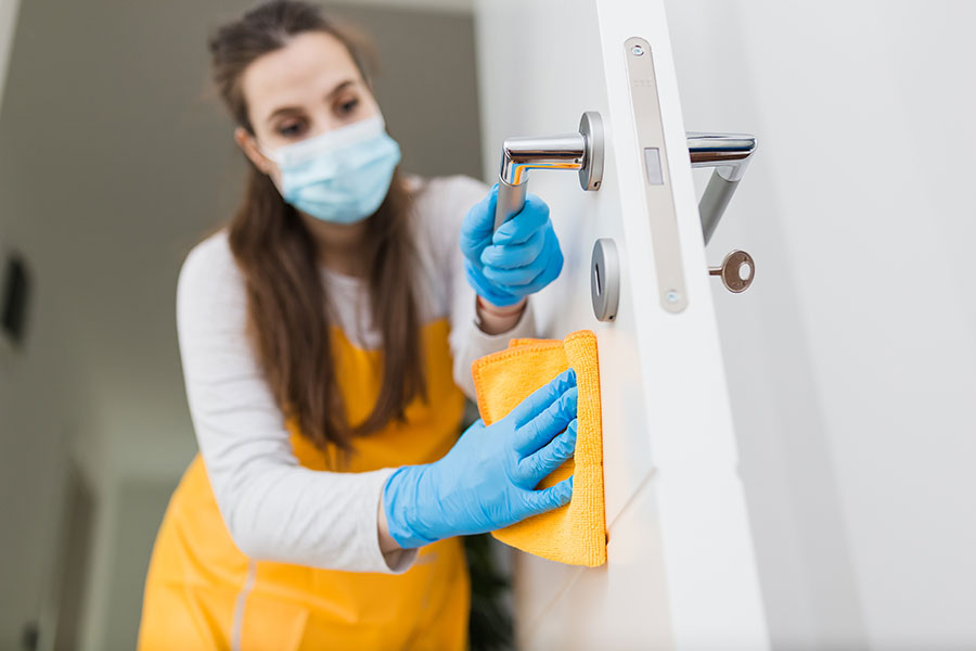 A professional maid in a long white sleeve shirt, yellow apron, and blue gloves wiping down a door handle in a small business in St. Charles, MO.