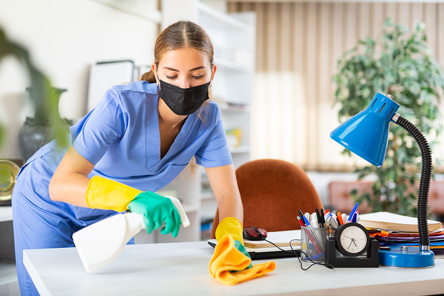 A young woman in blue scrubs and gloves uses a spray bottle and rag to clean an office desk in St Peters, MO.