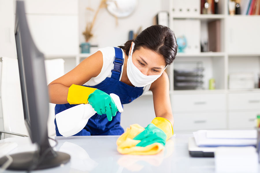 A professional cleaning lady in a white shirt and dark blue overalls is spraying a cleaning solution on an office desk in St. Peters, MO.