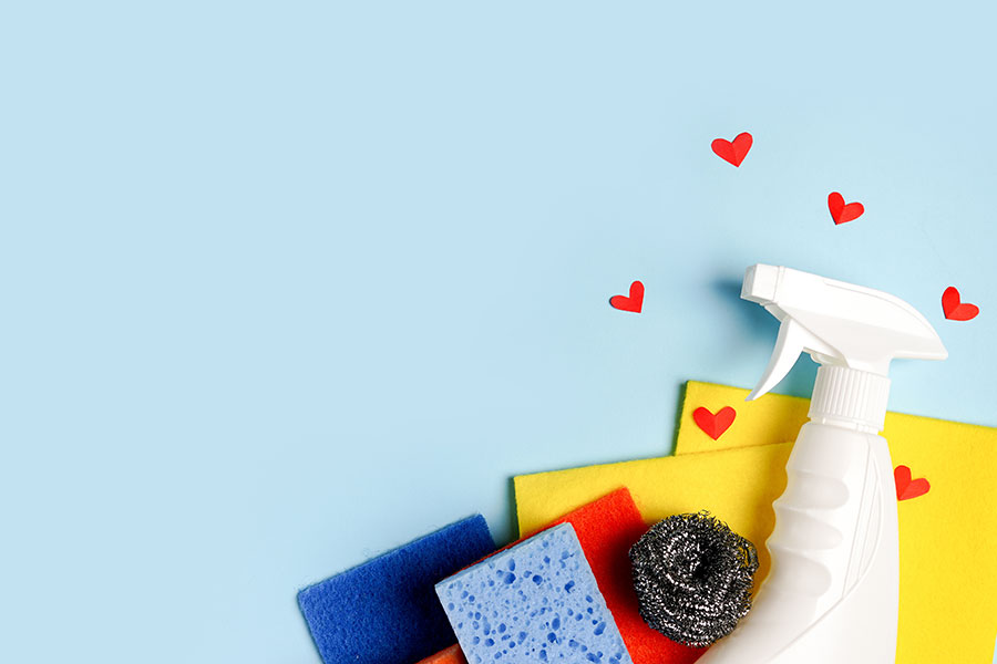 A pile of cleaning supplies against a blue background with hearts surrounding a white spray bottle for Valentine’s Day cleaning in Chesterfield, MO.