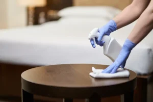 A woman wearing blue nylon nitrile gloves is cleaning a small bedside table in an apartment.